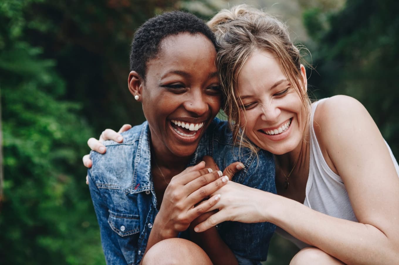 Two people laughing together outdoors among green foliage, leaning on one another.